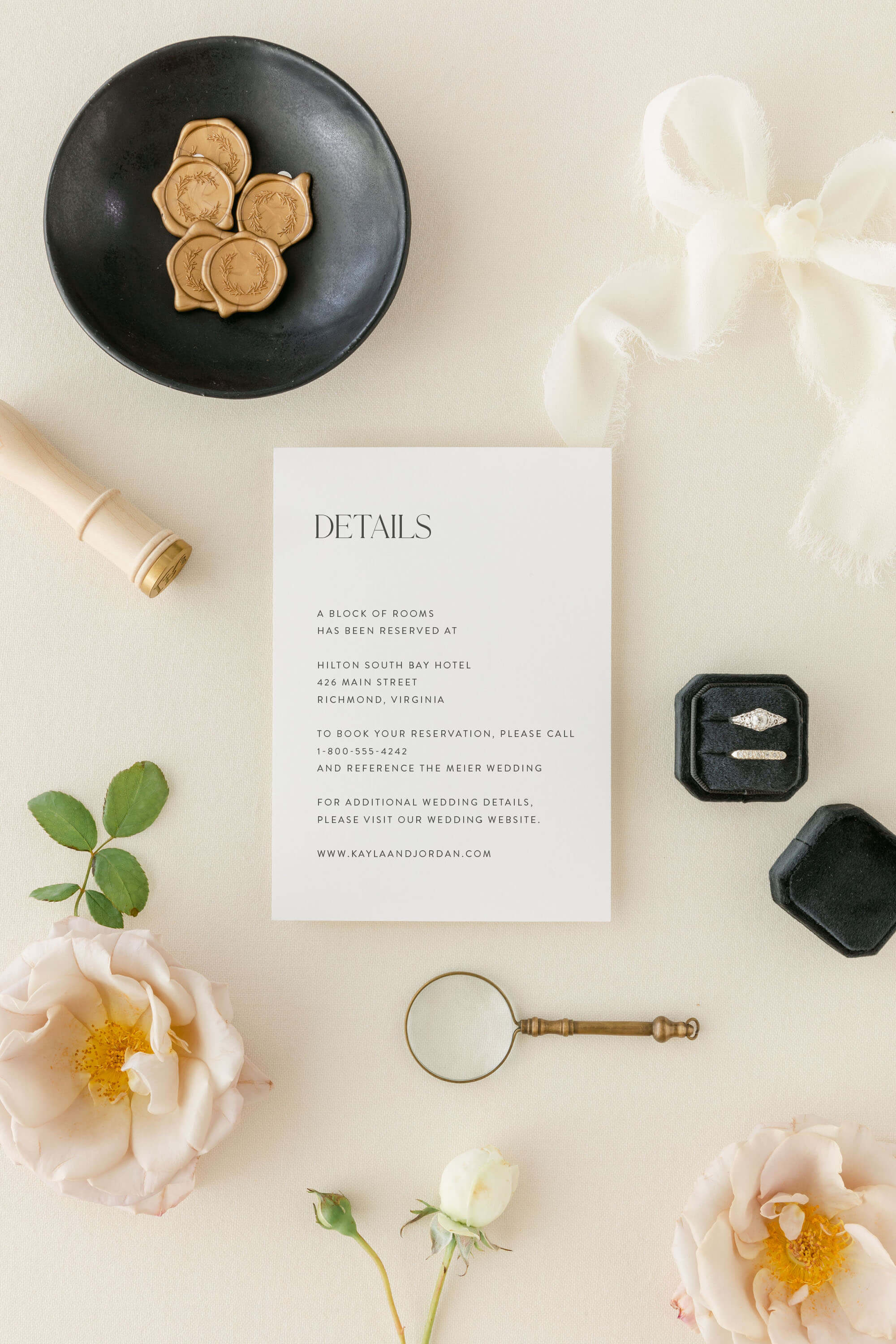 Flat lay with modern wedding details card, black plate with wax seals, white flowers, and decorative items on a light background.