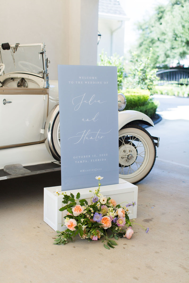 Dusty blue wedding welcome sign with white text on a white stand with peach flowers